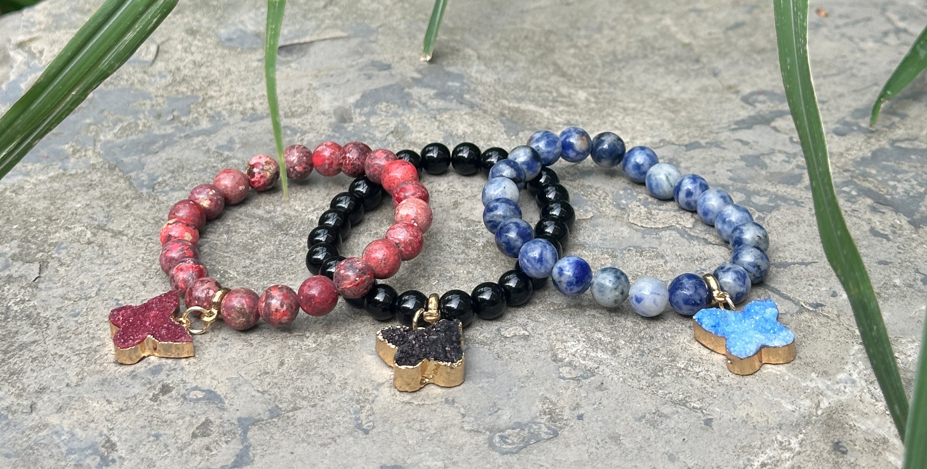 Three beaded stretch bracelets with star charms are arranged on a gray stone outdoors, surrounded by green grass. The bracelets feature red, black, and blue beads with gold-toned star charms.