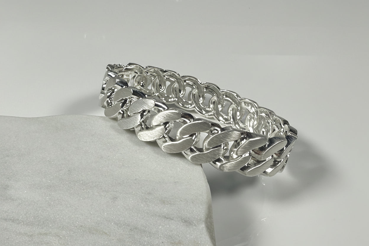 A silver-toned metal chain link bracelet with a wide curb-style link pattern, displayed on a light gray textured surface against a neutral light gray background.