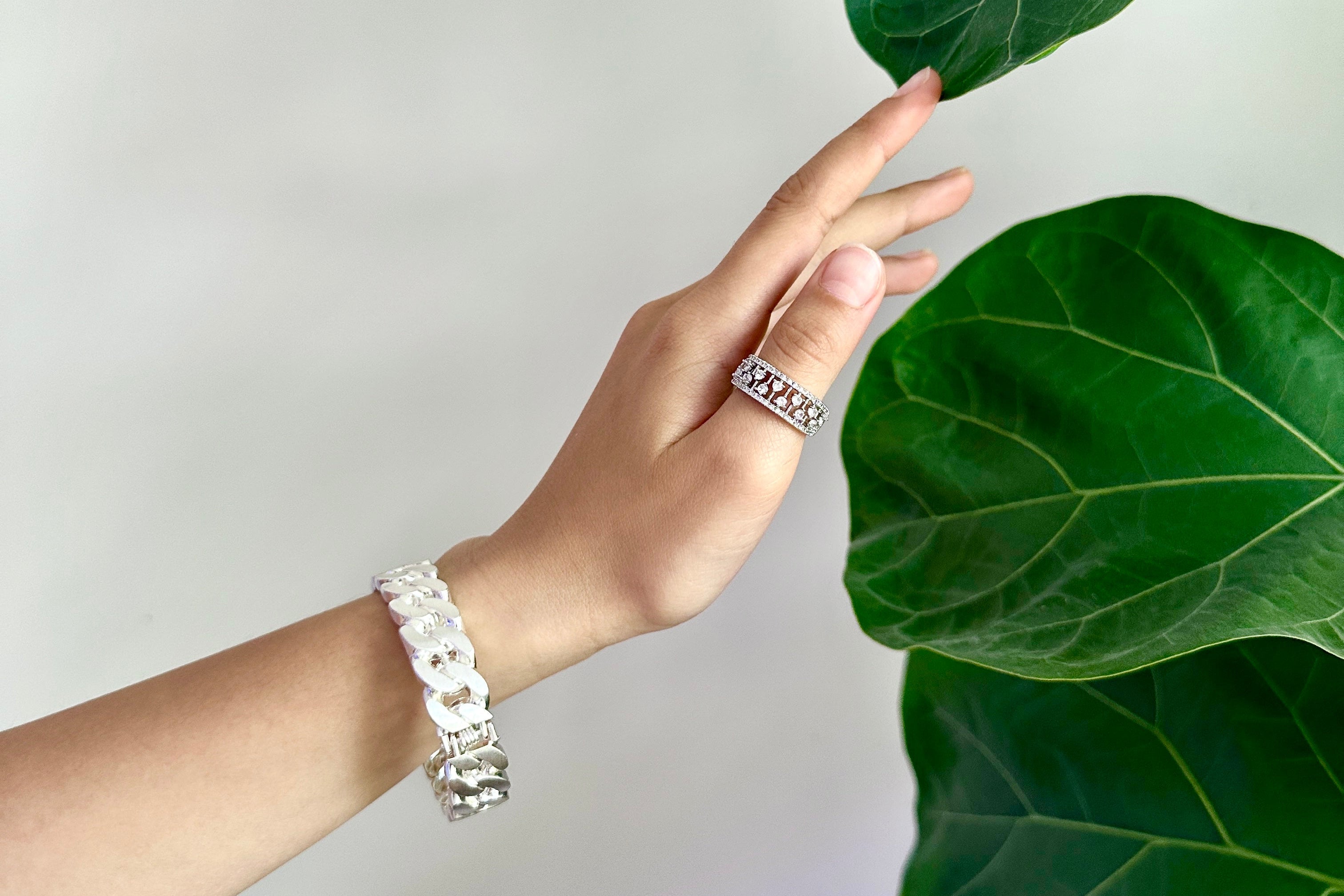 A person's hand wearing a silver ring and a white beaded bracelet, reaching toward large green fiddle-leaf fig plant leaves against a light gray background.